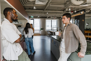 Two male employees lean on tables and stand facing each other while one of them talks and holds a cup