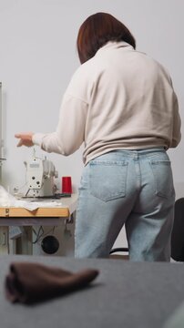 calm caucasian seamstress at sewing machine repairing garment, small atelier setting with denim, beige sweater, red thread spool on table, concentrated hands guiding fabric through industrial machine,