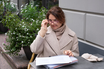 Woman with brown hair wearing a beige coat and glasses is seated at a table outdoors, reviewing documents with potted flowers in the background, showcasing a relaxed atmosphere