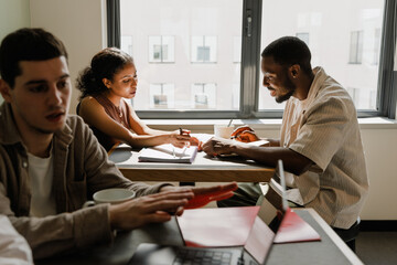 Fototapeta premium A male employee is talking and smiling while sitting at a desk across from a female employee and they are reading notebooks while a male employee is sitting next to them
