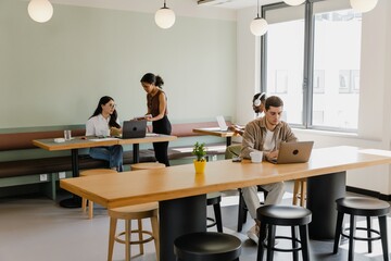 A female worker stands and shows a notebook to a female worker sitting at a desk while two male workers are typing on laptops nearby
