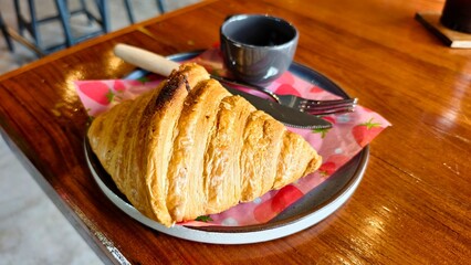 Golden croissant served on a plate with coffee and cutlery, resting on a strawberry-patterned napkin on a wooden cafe table.