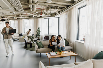 Obraz premium Two female employees are sitting on a couch reading a document while a male employee walks past them, looking at his phone and carrying a laptop