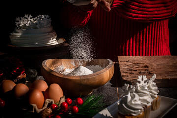 A person in a red knitted sweater sifts flour into a wooden bowl, creating a warm Christmas baking atmosphere with seasonal decorations and soft sunlight.