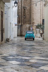 Obraz premium turquoise car driving away down a wet, narrow cobblestone alley in a historic southern italian town, flanked by whitewashed buildings and an ancient stone church on a moody day