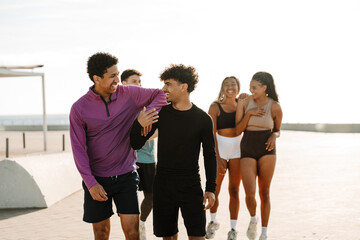 Obraz premium A male athlete rests his hand on the shoulder of a male athlete walking next to him while they laugh and behind them is a group of three athletes