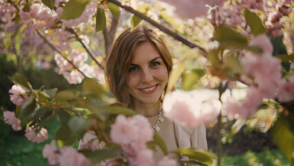 Fototapeta premium Smiling woman stands among blooming cherry blossoms in springtime park during daylight, enjoying nature and flowers while surrounded by pink petals