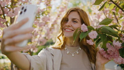 Young woman takes selfie among pink flowers in garden during bright day with sunlight shining on her face while smiling happily with her phone