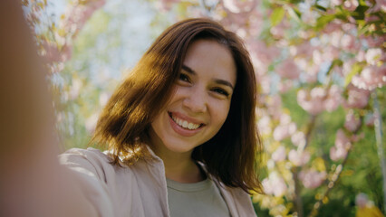 Young woman smiles while taking a selfie in a garden filled with blooming flowers during a sunny day in springtime