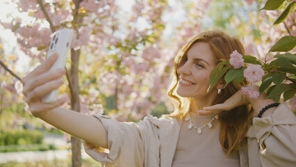 Woman takes selfie among blooming cherry trees in a park during spring afternoon