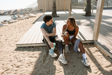 Male and female athletes talking and laughing while sitting, he holding a bottle and she holding a...
