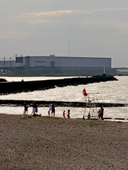People enjoying a sunny day at the beach with a large industrial building in the background