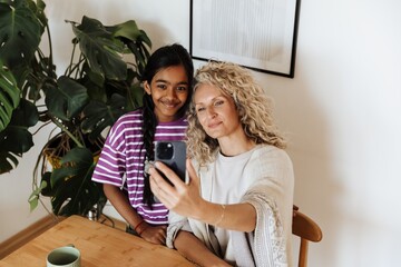 A mother sits at a table holding a phone and poses in front of it with her daughter standing next to her while they smile