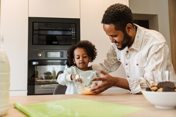 A daughter sits at a table and stirs a bowl with a fork while her father stands next to her and hugs her