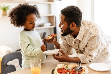 Father holding a slice of pizza and a plate while smiling and talking to his daughter while they stand at the table
