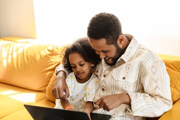 Obraz premium Father laughing and holding his arm around his daughter while they look at a laptop and sit on the couch