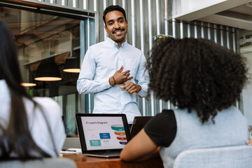 A male worker stands and smiles while listening to a female worker sitting across from him at a table next to a female coworker
