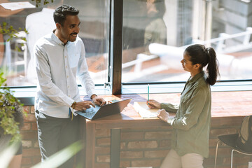 A male employee is talking to a female employee standing next to him at the desk and they are laughing