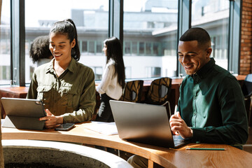 Female worker looking at computer tablet while male worker looking at laptop while they laugh at table