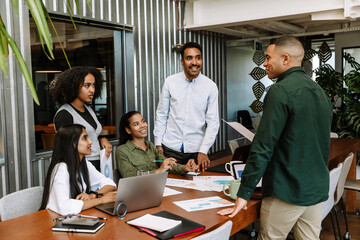 A male worker is talking to a group of four workers standing across from him and sitting at a table...