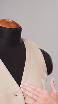 closeup white hands finishing beige vest on black mannequin, careful buttoning and loop adjustment, visible buttonhole and textured fabric, precision stitching and final inspection under soft studio