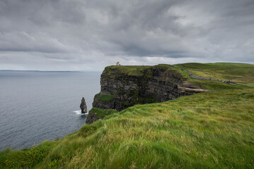 The Cliffs of Moher on Ireland&rsquo;s west coast, with green grass leading to the cliff edge under a grey sky, featuring a stairway with tourists, showcasing the natural landscape and geological features