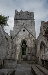 The stone Muckross Abbey in Ireland with a prominent tower and arched entrance under an overcast sky