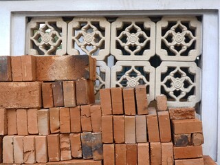 A messy pile of raw, reddish-orange clay bricks stacked in the foreground, set against a blurred background of a traditional decorative off-white floral ventilation screen.