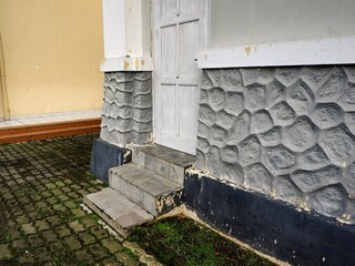 Weathered entrance of an aged building. Steps lead to an old wooden door, framed by a textured, decorative concrete foundation and overgrown pathway.