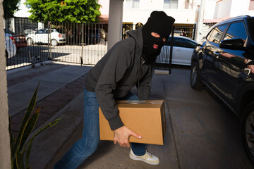 Masked porch pirate stealing a delivery package from a residential home, covering face with a ski...