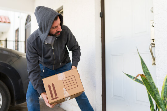 Man in a hoodie furtively stealing a new delivery package from a residential doorway. Concept of property crime and theft