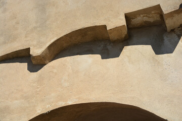 A close-up of a weathered, sandy-colored plaster wall features a curving, stepped architectural...