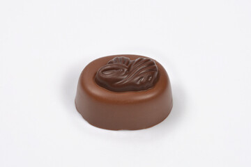 A single, cylindrical milk chocolate praline with a raised, dark chocolate swirl design on top is centered on a plain white background