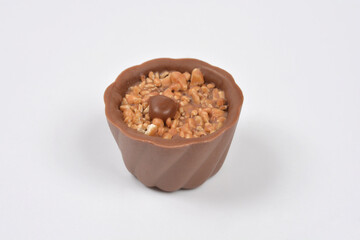 A single, chocolate cup praline is viewed from the side, topped with chopped hazelnuts and a whole chocolate-covered hazelnut on a white background