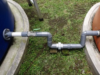 Overhead view of a gray PVC pipe with a green ball valve connecting the base of a blue water tank to an orange water tank, set on a grassy area with concrete curbs.