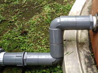 A clean, close-up view of an L-shaped gray PVC pipe and coupling connecting to the side of a large orange plastic water storage tank, resting near a concrete curb.