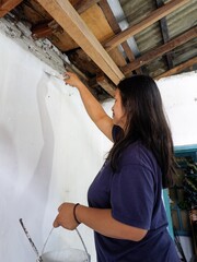 A woman uses a brush to apply white paint to a textured interior wall, working near the ceiling to cover peeled plaster and exposed wooden roof beams during home renovation.