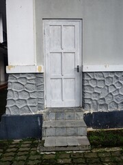 A quiet, weathered white wooden six-panel door framed by gray walls and a decorative stone-patterned foundation. Low stone steps lead up from mossy brick paving.