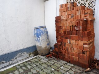 A large, neat stack of red clay bricks is stored in a corner next to a dirty, battered blue plastic barrel and moss-covered interlocking concrete pavers.