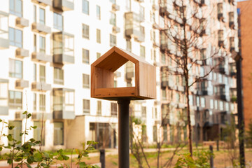 Wooden stylized bird feeder in form of small house against backdrop of modern apartment buildings, symbolizing care for birds and community living. Affordable housing, mortgages