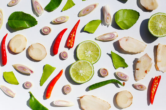 A colorful, close-up pattern of Thai herbs: lime slices, galangal, lemongrass, chilies, and kaffir lime leaves.