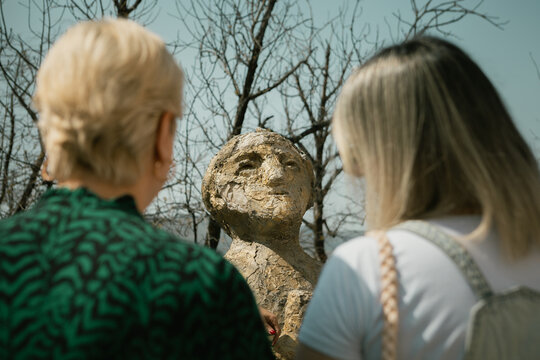 Sighnaghi, Georgia - 3 29 2025: Two women admire a weathered statue of Tamada, the Georgian Toastmaster, under a bright sky with bare trees in the background.