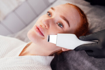 Red-haired woman receiving facial treatment with ultrasonic device in a spa setting, showcasing skincare process and relaxation in a serene environment