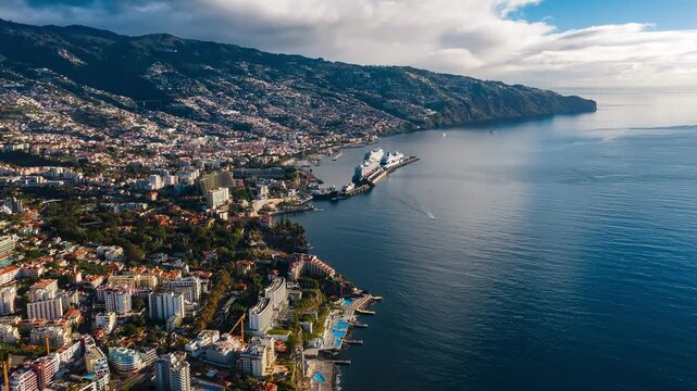 Aerial view of Funchal, Madeira shows harbor with two cruise ships, long pier, cliffside hotels, pools, and volcanic cliffs in late day light with calm water reflections.