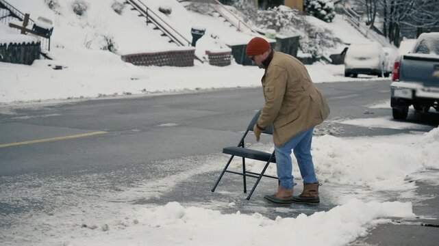 A man sits in a folding chair in a cleared street parking space after shoveling snow, symbolizing the Pittsburgh tradition of saving parking spots. 14978  