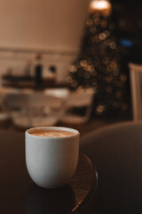 A cozy, warm scene of a cup of cappuccino in a modern cafe during the holiday season The blurred Christmas tree in the background adds a festive touch