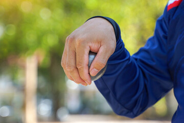 A person's hand holds a silver metal pétanque (bocce) boule (ball) in preparation for a throw,...