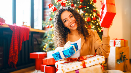 A curly-haired woman with a sparkler and Christmas presents sits near a decorated tree in a cozy...