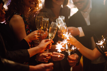 Young friends having fun with sparklers and glasses of champagne at corporate party. Handsome men and woman spend time together, celebrating New Year in cozy space. Сoncept is festive and fun.