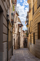 Naklejka premium Vertical view down a narrow cobblestone Toledo street, lined with warm stucco facades, wrought-iron balconies, exposed stone, and a covered wooden balcony in the distance
